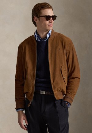 Brun bomberjakke i ruskind med ribstrikkede manchetter og talje. Båret over en marineblå sweater og mønstret skjorte, suppleret med solbriller.