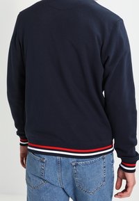 Marineblauwe sweater met een soepele textuur, voorzien van een geribde zoom met rode en witte strepen. Lichtblauwe denim jeans met twee achterzakken.