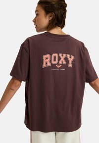 Žena v volném tmavě fialovém tričku s potiskem "ROXY" a "Paradise Found" na zádech, stojící s mírně nataženýma rukama.