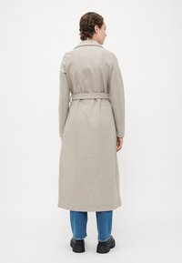 Persona che indossa un lungo cappotto beige con cintura, jeans blu e scarpe nere spesse, in piedi di spalle su uno sfondo bianco puro.