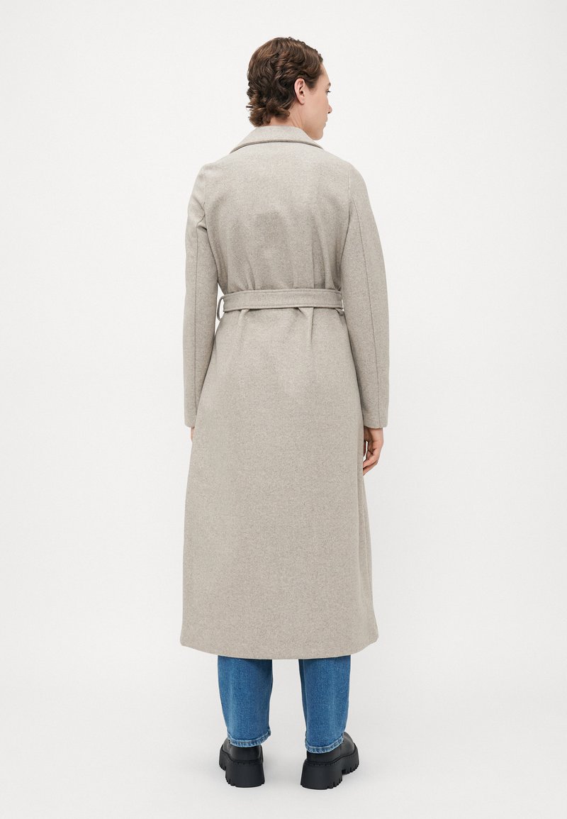 Personne portant un long manteau beige ceinturé, un jean bleu et des chaussures noires massives, se tenant de dos devant un fond blanc uni.