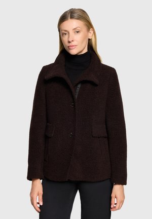 Donna che indossa un cappotto marrone scuro con texture, collo e bottoni sopra un dolcevita nero, in piedi davanti a uno sfondo chiaro e uniforme.