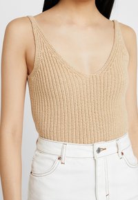 Beige neule toppi syvällä V-päällä ja ohuilla olkaimilla, jossa on teksturoitu kangas. Yhdistetty korkeavyötäröisiin valkoisiin farkkushortseihin.