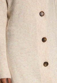 Pitkä neuletakki pehmeästä, kermaisen värisestä villasekoitteesta, jossa hieman teksturoitu viimeistely. Mallissa neljä pyöreää ruskeaa nappia edessä.