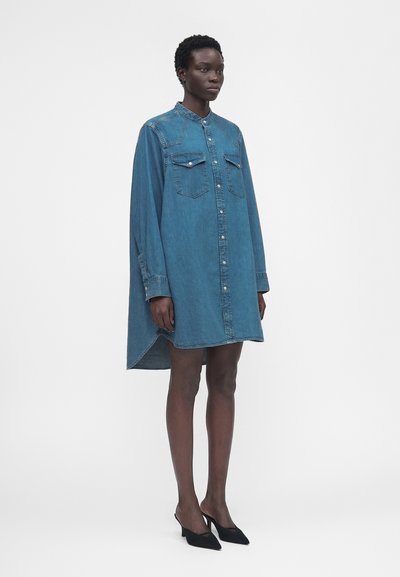 Γυναίκα στέκεται φορώντας ένα μπλε ντένιμ φόρεμα πουκάμισο, άνετο και φαρδύ, μήκους μέχρι το γόνατο, με τσέπες μπροστά και μαύρα μυτερά τακούνια σε λευκό φόντο.