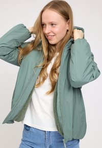 Hellgrüne Jacke mit einem weichen, strukturierten Innenfutter. Verfügt über einen Reißverschluss an der Vorderseite, eine Kapuze und elastische Bündchen. Trägt man über einem weißen Hemd und blauen Jeans.