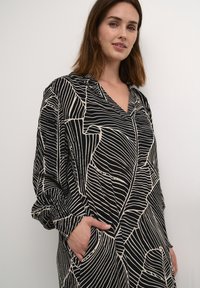 Zwarte en crème gestreepte jurk met een V-hals, lange mouwen en zakken. Bevat abstracte lijn ontwerpen en heeft een losse pasvorm.