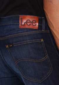 Jeans de mezclilla azul oscuro con un parche de cuero que presenta el logo de 'Lee'. Costuras visibles en amarillo y diseño clásico de cinco bolsillos.