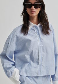 Camicia a righe blu con colletto bianco, caratterizzata da una vestibilità rilassata e una texture morbida. Dettagli con bottoni sulle maniche.