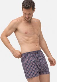 Hombre sin camiseta ajustándose unos calzoncillos a cuadros azules y rojos contra un fondo blanco liso.