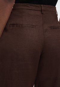 Κοντινό πλάνο ατόμου που φοράει σκούρο καφέ υφασμάτινο παντελόνι με τσέπες welt στην πίσω πλευρά και μαύρο τοπ μέσα στο παντελόνι.