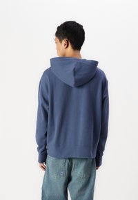 Μπλε βαμβακερή μπλούζα με μεγάλη κουκούλα, ριγέ ώμους και πλεκτούς μανίκες. Φοριέται με ανοιχτό μπλε τζιν, που διαθέτουν πίσω τσέπες.