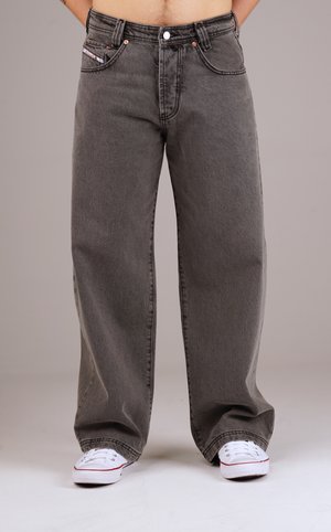 Person wearing loose gray denim jeans and white sneakers, standing against a plain gray background with hands in pockets.