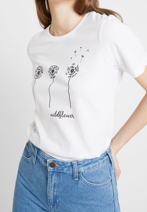 Valkoinen t-paita, jossa mustat voikukkakuvitukset ja teksti "wildflower", pidetty sinisten korkeavyötäröisten farkkujen kanssa, käsi lepää lantion päällä.