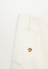 Tissu en denim blanc avec un rabat de poche rectangulaire maintenu par un seul bouton marron, montrant des coutures détaillées sur un fond clair.