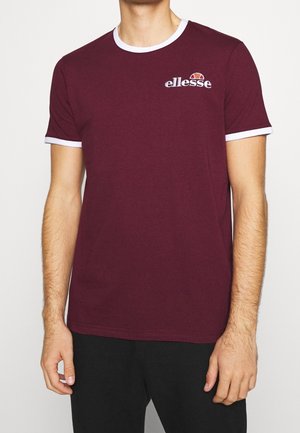 Mand iført en kirsebærrød Ellesse t-shirt med hvide kanter ved krave og ærmer, kombineret med sorte bukser, stående foran en ensfarvet baggrund.
