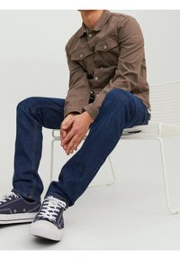 Hnědá denimová bunda s kovovými knoflíky, v kombinaci s tmavě modrými džíny a námořnickými teniskami s bílými tkaničkami, sedí na bílé židli.