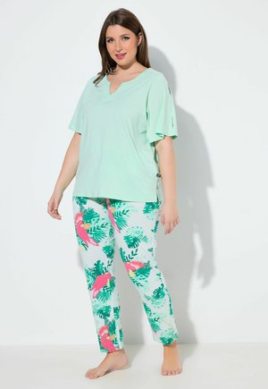 Kobieta stoi boso, ubrana w miętowozielony krótki rękaw i białe spodnie z wzorem różowego papugi i zielonych liści.