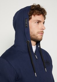 Dunkelblaue Kapuzenjacke mit durchgehendem Reißverschluss, ausgestattet mit schwarzen Kordeln und einem Logo-Patch. Glattes Material mit schlichtem Design und einer strukturierten Kapuze.