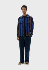 Gestreepte jas in blauw, bruin en zwart met een kraag, knoopsluiting en twee klepzakken. Gecombineerd met donkere spijkerbroek en casual schoenen.