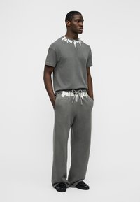 Man standing with hands in pockets wearing gray short-sleeve shirt and matching loose pants with white text accents and black shoes.