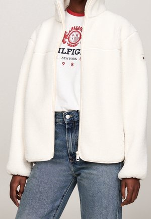 Person iført en uåbnet cremefarvet fleecejakke, hvid t-shirt med røde detaljer og blå højtaljede jeans, som står med afslappede arme.