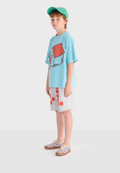 Garçon portant une casquette turquoise, un t-shirt bleu avec un motif de maison rouge, un short gris avec des carrés rouges, et des baskets rayées, debout de profil.