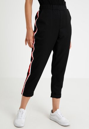Person trägt schwarze kurze Hosen mit roten und weißen Seitenstreifen und weiße Sneakers und steht vor einem einfarbigen Hintergrund.