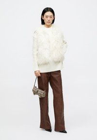 Colete de pelagem sintética branca sobre um suéter creme, combinado com calças boca de sino castanhas. Modelo segura uma mala metálica. Design simples e texturizado.