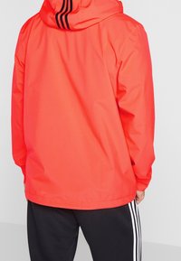 Red waterproof jacket with a hood, featuring black stripes on the back, smooth texture, and long sleeves, paired with black pants.