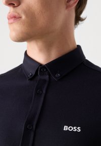 Marineblå polo skjorte med knapp-ned krage, teksturert stoff og hvit brodert "BOSS" logo på brystet. Knapper er mørke og runde.