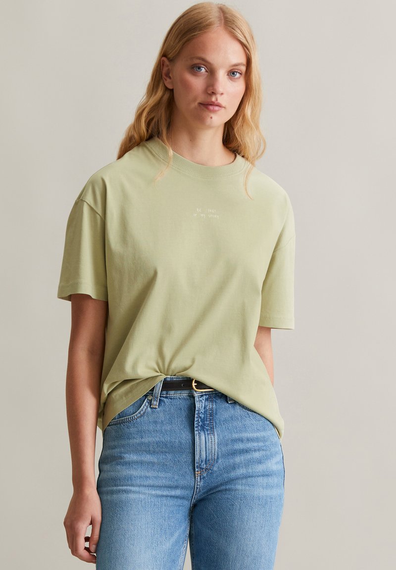 T-shirt oversized verde chiaro con maniche corte e scollatura tonda. Testo "Fai parte della mia storia" stampato in bianco sulla parte anteriore.