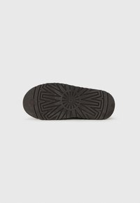 Rubberen zool van een donkerbruine UGG-schoen met geometrisch profiel en een cirkelvormig logo in het midden op een eenvoudige witte achtergrond.