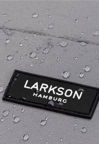 Grått tyg med vattendroppar, med en svart rektangulär etikett som har "LARKSON HAMBURG" i vitt text. Texturerad ytdetalj synlig.