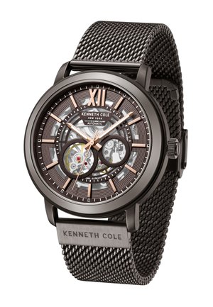 Montre avec bracelet en maille noir, mouvement automatique transparent, accents en bronze et un chiffre XII bien en évidence. Présente un cadran texturé et un logo.