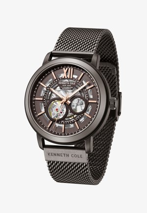 Black mesh strap watch with transparent automatic movement, bronze accents, and prominent XII numeral. Features textured dial and branding.
