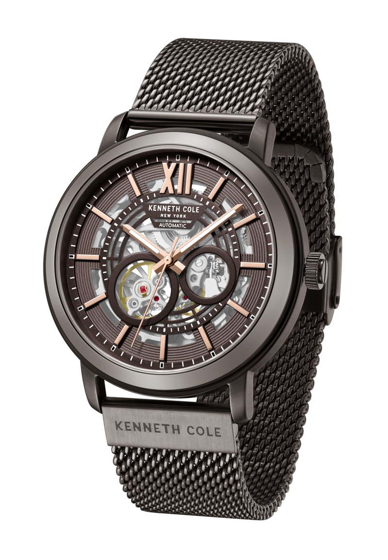 Montre avec bracelet en maille noir, mouvement automatique transparent, accents en bronze et un chiffre XII bien en évidence. Présente un cadran texturé et un logo.