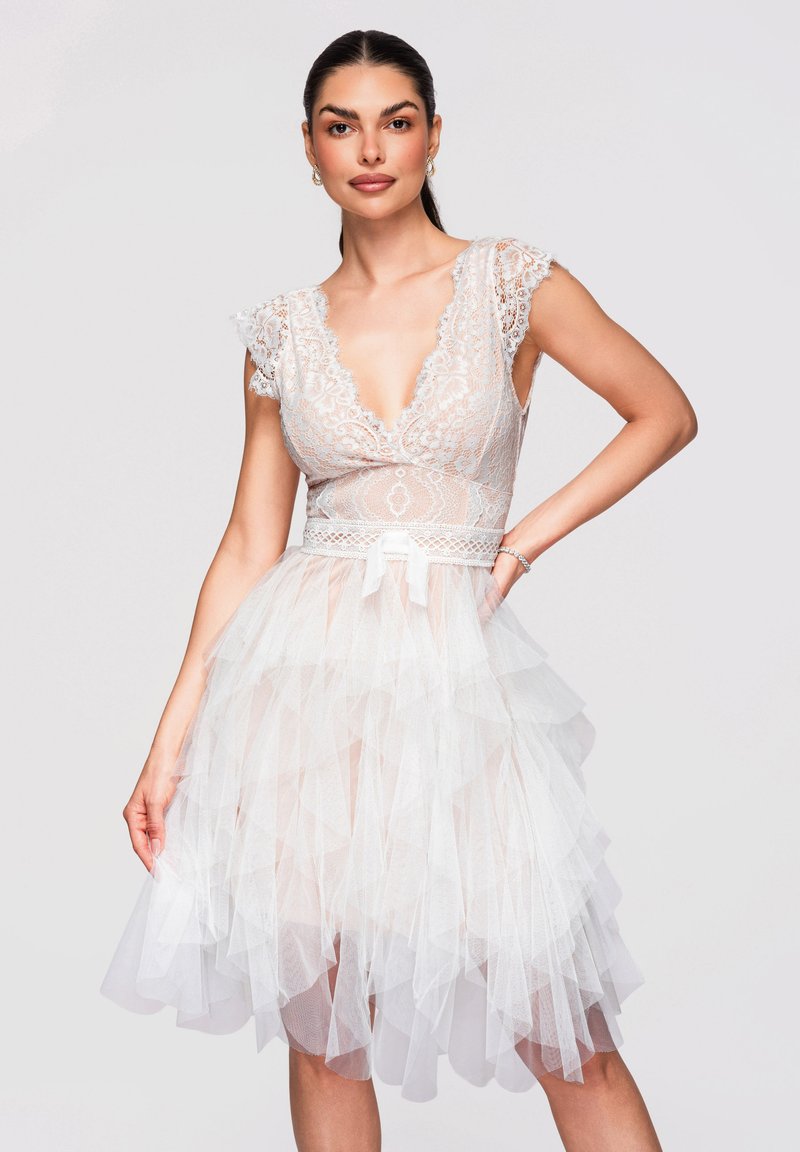 Femme portant une robe en dentelle blanche avec un décolleté en V profond, une jupe en tulle froufroutante et des manches courtes, posant avec une main sur la hanche devant un fond uni.