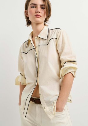 Eine junge Person trägt ein cremefarbenes Langarmshirt mit schwarzer Paspelierung und cremefarbene Hosen mit braunem Gürtel und steht vor einem schlichten hellen Hintergrund.