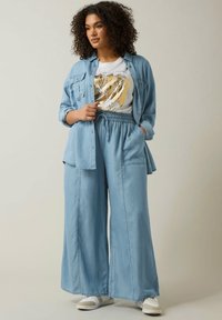 Camicia chambray azzurro chiaro sopra una t-shirt bianca con dettagli dorati, abbinata a pantaloni a gamba larga azzurro chiaro e sneaker bianche.