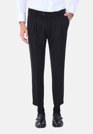 Pantaloni neri su misura con fronte piatto, pieghe e tasche, realizzati in un tessuto liscio. Indossati con scarpe e calze nere.