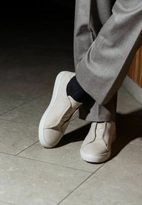 Beige suede sneakers with elastic side panels, white rubber soles, and a textured finish, paired with gray tailored pants and dark socks.