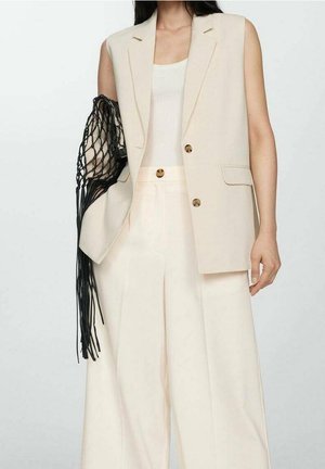 Woman wearing sleeveless beige blazer, matching wide-leg trousers, cream tank top, and holding black netted fringe bag on arm.