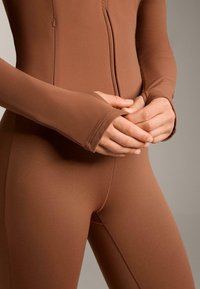 Completo marrone realizzato in tessuto liscio e elasticizzato, con zip frontale e maniche lunghe con foro per il pollice. Primo piano delle mani sui fianchi.
