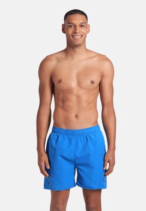 Giovane uomo che sorride, in piedi, con pantaloni da bagno blu brillante, senza maglietta, su uno sfondo bianco uniforme.