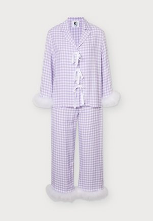Femme portant un ensemble de pyjama à carreaux violet clair et blanc avec des poignets et un ourlet blancs et duveteux, debout devant un fond clair uni.