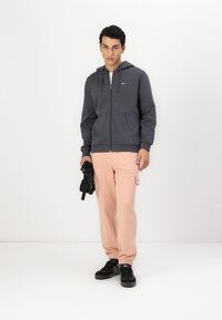 Grå zip-up hettegenser med store lommer, kombinert med lys rosa cargo bukser med sidelomme, og svarte joggesko. Enkelt design med fargekontrast.