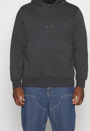 Hombre que lleva una sudadera negra con bolsillo frontal y vaqueros azules, de pie frente a un fondo liso y claro.