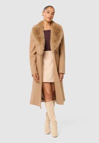 Beige ullkappa med en bred krage i konstpäls, dubbelknäppta knappar och en lång siluett, bärs över en ljus kjol och knähöga stövlar.