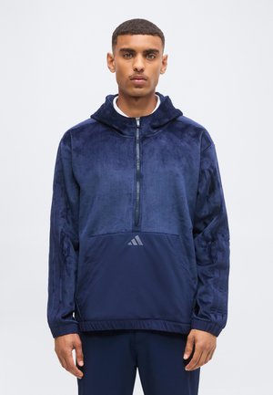 Mladý muž nosí tmavomodrý pulóver s kapucňou a štvrťovým zipsom, s predným vreckom a logom Adidas, stojí pred jednoduchým svetlým pozadím.